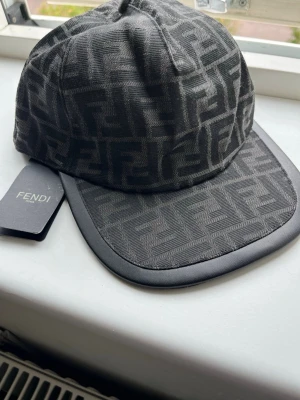 Svart keps med Fendi-logga - Snygg svart keps från Fendi med det klassiska FF-monogrammet över hela kepsen. Kepsen har böjd skärm med svart kant och justerbar rem bak. Tillverkad i tyg med exklusiv känsla och tydlig logodetalj.