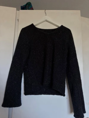 Svart glittrig stickad tröja - Mysig svart stickad tröja med glittertrådar som ger ett snyggt skimmer. Tröjan har rund halsringning, långa ärmar och lätt utsvängda ärmslut. Perfekt för dig som vill ha något extra till din outfit.