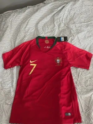 Portugal matchtröja Ronaldo #7 - Säljer en röd Portugal matchtröja från Nike med guldiga detaljer, nummer 7 och Ronaldo på ryggen. Tröjan har korta ärmar, gröna detaljer vid kragen och Portugals landslagsemblem på bröstet. Perfekt för dig som älskar fotboll och vill stötta landslaget. Helt ny aldrig använd. Säljer då den va för stor! 