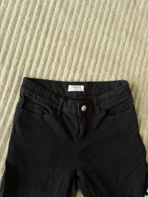 Svarta jeans från Lindex - Snygga svarta jeans från Lindex med klassisk femficksmodell och knappgylf. Jeansen har en rak passform och är tillverkade i ett mjukt bomullsmaterial med lite stretch för extra komfort.