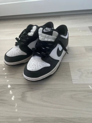 Nike Dunk Low svart/vit sneakers - Säljer ett par Nike Dunk Low sneakers i svart och vitt. Skorna har klassisk låg siluett, svart swoosh och vita partier i läder. Perforerad tåbox för extra ventilation och svarta skosnören. Perfekta för dig som gillar streetwear och tidlös stil. 