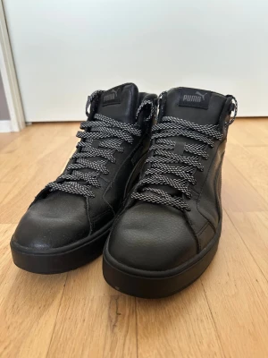 Svarta Puma high top sneakers - Snygga svarta high top sneakers från Puma med svart och vita mönstrade skosnören. Skorna är tillverkade i läder med klassisk Puma-logga på plösen och sidan. Tjock sula och stilren design som passar till många olika outfits.