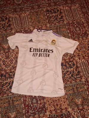 Real Madrid matchtröja Vinicius Jr size XL - Säljer en vit Real Madrid matchtröja från Adidas med tryck 'Emirates Fly Better' på framsidan och 'VIN JR 20' på ryggen. Tröjan har klubbmärke, korta ärmar och lila detaljer vid kragen. Perfekt för dig som älskar fotboll och vill stötta ditt lag. Size XL Old edition.