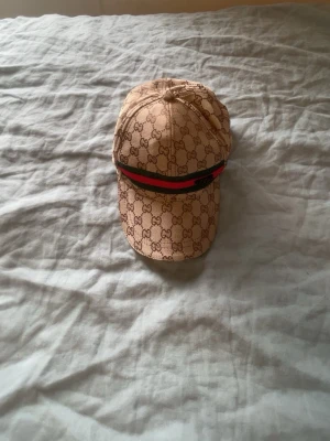 Beige keps med mönster! - Säljer en beige keps från  med det klassiska mönster monogrammet över hela kepsen. Kepsen har en böjd skärm och en ikonisk grön och röd rand framtill. Perfekt för dig som vill ha en lyxig och trendig accessoar till din outfit.