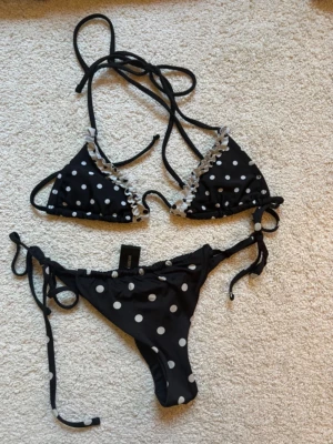 Svart bikini med vita prickar - Snygg svart bikini med vita prickar och volangdetaljer på överdelen. Klassisk trekantsmodell med knyt både i nacken och ryggen samt på sidorna av underdelen. Perfekt för strandhäng eller poolparty!