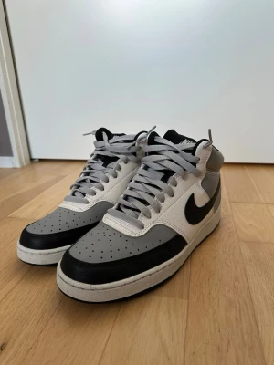 Nike grå och svarta sneakers - Snygga sneakers från Nike i grått, svart och vitt. Skorna har klassisk swoosh på sidan, perforerad tåbox och grå skosnören. Ovandelen är i läder och designen är tidlös med en sportig vibe. Perfekta för dig som gillar streetwear och vill ha något stilrent.