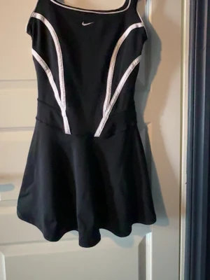 Svart sportklänning från Nike -  Svart tennis  klänning från Nike med vita detaljer längs sidorna och axelbanden. Den är  stretchig och har en inbyggd sport BH