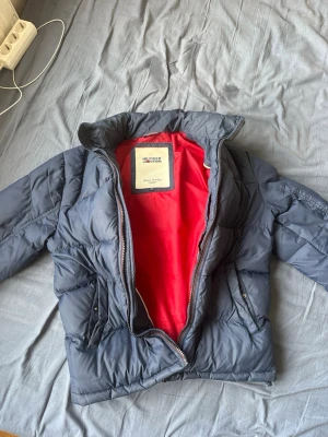 Mörkblå pufferjacka från Tommy Hilfiger - Snygg mörkblå pufferjacka från Tommy Hilfiger med röd insida. Jackan har hög krage, dragkedja framtill och två fickor med tryckknappar. Klassisk quiltad design och diskret logga på ärmen. Perfekt för kalla dagar.