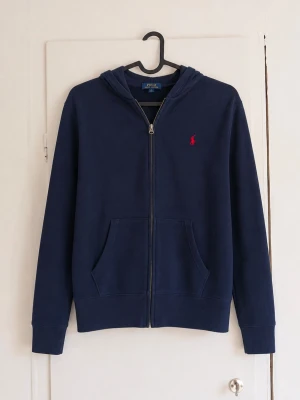 Ralph Lauren zip hoodie - Klassisk mörkblå zip hoodie från Polo Ralph Lauren med broderad röd logga på bröstet. Stilren och tidlös modell med dragkedja, huva och framfickor – perfekt till både vardag och höst/vår. ✓ Original Polo Ralph Lauren ✓ Mörkblå färg med röd broderad logga ✓ Dragkedja och huva ✓ Två framfickor ✓ Bekväm passform ✓ Bra skick men har några defekter!
