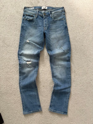 Blå slitna jeans från Jack & Jones - Snygga blå jeans från Jack & Jones i modellen Slim Glenn. Jeansen har slitningar och hål på låret och knät för en trendig look. Klassisk femficksmodell med dragkedjegylf och bälteshällor. Perfekta för en avslappnad stil.