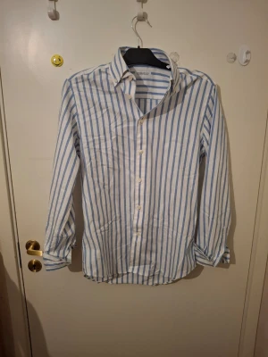 Randig skjorta från These Glory Days - Säljer en klassisk skjorta från These Glory Days med vita och blå vertikala ränder. Skjortan har lång ärm, knappar framtill och en avslappnad passform. Perfekt till jeans eller chinos för en clean look.