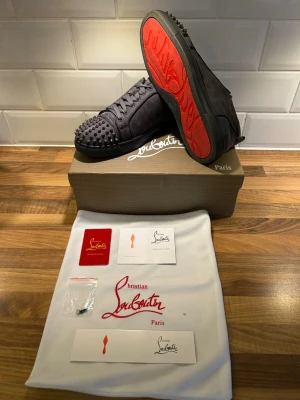 Christian Louboutin grå spikes sneakers - Grå sneakers från Christian Louboutin i mocka med ikoniska röda sulor och grå nitar över tån. Skorna har klassisk låg profil och snörning. Kommer med originalkartong, dustbag och certifikat. Perfekta för dig som vill sticka ut med exklusiv stil.