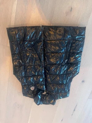 Svart glansig dunväst med dragkedjor från Moncler! - Snygg svart dunväst med glansigt material och två framfickor med dragkedja. Västen har hög krage och quiltad design som ger en modern look. Perfekt för lager-på-lager-stil under kyliga dagar.