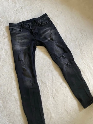 Svarta DSQUARED2 jeans - Säljer ett par svarta DSQUARED2 jeans med snygga slitningar och tvättade detaljer på benen. Jeansen har fem fickor och knappgylf. Perfekta för dig som gillar en trendig och avslappnad stil.