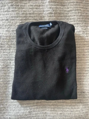 Svart tröja från Polo Ralph Lauren - Klassisk svart tröja från Polo Ralph Lauren med rund halsringning och diskret lila logga broderad på bröstet. Tröjan har en enkel och stilren design, perfekt att matcha med jeans eller chinos. Materialet känns mjukt och bekvämt.