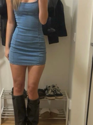 Blå tight jeansklänning - Säljer en blå tight jeansklänning med smala axelband och kort längd. Klänningen har en figurnära passform och markerade sömmar som ger en snygg siluett. Perfekt för dig som gillar en enkel men trendig look.