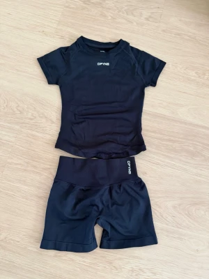 Svart träningsset från DYNE - Säljer ett svart träningsset från DYNE med kortärmad topp och matchande shorts. Setet är gjort i ett stretchigt, snabbtorkande material som passar perfekt för gym eller löpning. Både tröjan och shortsen har logga framtill. Snygg och sportig look!