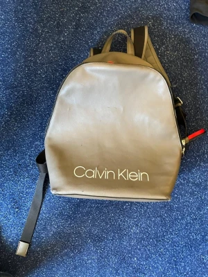 Grå ryggsäck från Calvin Klein - Snygg grå ryggsäck från Calvin Klein i slätt konstläder. Väskan har ett stort huvudfack, svarta justerbara axelband och en cool detalj med röd och vit rand längs dragkedjan. Perfekt för dig som vill ha en stilren och trendig accessoar.