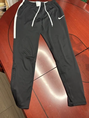 Svarta Nike mjukisbyxor med vit detalj - Snygga svarta mjukisbyxor från Nike med vit dragsko i midjan och vit rand längs sidan. Byxorna har smal passform och är perfekta för chill eller träning. Klassisk Nike-logga på vänster ben och elastisk midja för skön känsla.