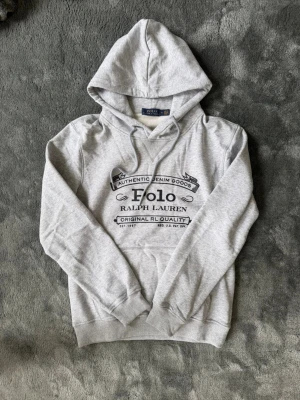 Grå hoodie från Polo Ralph Lauren - Snygg grå hoodie från Polo Ralph Lauren med svart tryck framtill där det står 'Authentic Denim Goods' och 'Original RL Quality'. Klassisk huva med dragsko och mjukt material, perfekt för en chill och avslappnad stil.