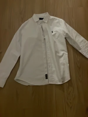 Vit skjorta från Ralph Lauren - Klassisk vit skjorta från Ralph Lauren med lång ärm och knappar framtill. Skjortan har den ikoniska loggan broderad på bröstet och är tillverkad i bomull för en skön och stilren look. Perfekt till jeans 