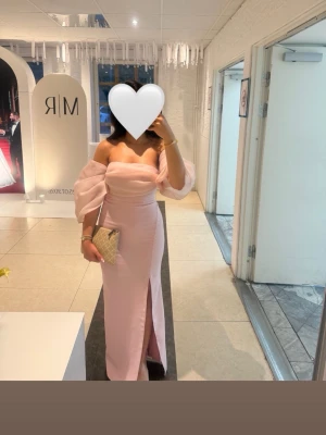 Ljusrosa långklänning  - Elegant ljusrosa långklänning med offshoulder-design och puffiga ärmar. Klänningen har en slits framtill och figurnära passform. Endast använd 1 kväll. Passar XS/S