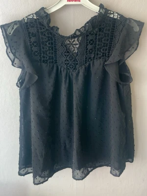 Svart blus med spets och volang - Snygg svart blus med transparenta prickiga detaljer och spets upptill. Blusen har volang vid ärmslut och halsringning samt korta ärmar. Perfekt för dig som gillar en romantisk och trendig stil.
