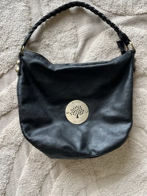 Svart handväska i läder - Snygg svart handväska i mjukt läder med flätat handtag och guldfärgade metalldetaljer. Väskan har en rund form och ett stort emblem framtill. Dragkedja upptill och innerfack för småsaker. Sliten i både handtaget och lite på innersidorna men inget man ser
