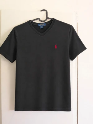 Polo Ralph Lauren svart T-shirt – M (10–12 år) - Svart T-shirt från Polo Ralph Lauren med klassisk röd broderad logga på bröstet 🐎 V-ringad modell och enkel, stilren design som passar till det mesta. Storlek: M / 10–12 år Färg: Svart med röd logga Skick: Fint begagnat skick, inga defekter förutom att den är lite färg blekt! Därav priset😁 Made in Vietnam. Säljer då den inte längre kommer till användning. Hör gärna av dig vid frågor eller om du vill ha fler bilder! 😊