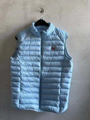 Ljusblå dunväst från Jott - Snygg ljusblå dunväst med ståkrage och quiltad design. Västen har dragkedja framtill och ett diskret märke på bröstet. Perfekt att slänga över en hoodie för en chill och trendig look. Passar till både jeans och mjukisbyxor.