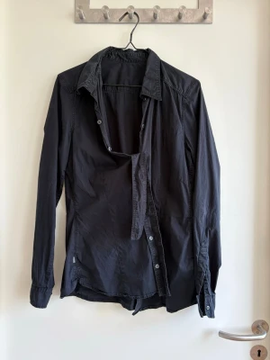 Svart långärmad skjorta - Stilren svart skjorta med klassisk krage och långa ärmar. Skjortan har knappar framtill och är tillverkad i ett mjukt bomullstyg. Perfekt för dig som gillar en enkel och clean look.