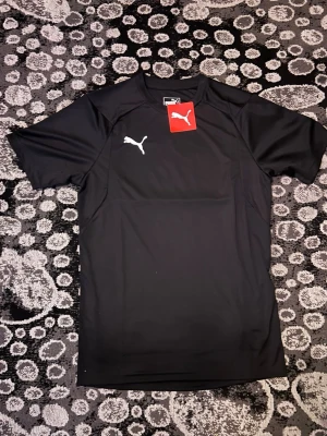 Svart träningströja från Puma - Säljer en svart träningströja från Puma i storlek XS. Tröjan har korta ärmar och en vit Puma-logga på bröstet. Perfekt för träning eller sportaktiviteter. Tillverkad i ett lätt och funktionellt material som andas.