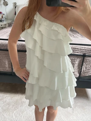 Vit one shoulder volangklänning - Säljer en vit kortklänning med one shoulder-design och flera lager volanger. Klänningen har en lös passform och är perfekt för dig som gillar en romantisk och trendig stil. Tillverkad i ett lätt tyg som ger ett snyggt fall.