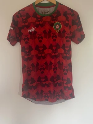 Marocko matchtröja från Puma - Säljer en officiell Marocko matchtröja från Puma i storlek S. Tröjan har ett rött och svart geometriskt mönster, grönt runt kragen och Marockos fotbollsförbunds märke på bröstet. Tillverkad i lätt och ventilerande material, perfekt för träning eller match.