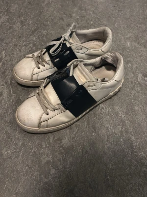 Vita sneakers med svart band - Säljer ett par vita sneakers med breda svarta band över vristen och gråa skosnören. Skorna har en klassisk låg profil och är tillverkade i slätt lädermaterial. Perfekta för en clean och stilren look.
