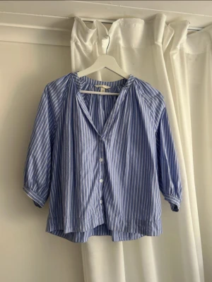Blåvit randig blus med knappar - Snygg blå blus med vita vertikala ränder och knäppning framtill. Blusen har lös passform, trekvartsärmar och en lätt v-ringad halsringning. Perfekt för en avslappnad och trendig look.