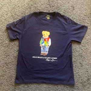  Ralph Lauren shirt - Säljer en mörkblå t-shirt från Ralph Lauren med den ikoniska Polo Bear-trycket på bröstet. T-shirten har korta ärmar, rund hals och är i en slim fit-modell. Perfekt för dig som gillar klassisk men lekfull stil. Priset kan diskuteras 