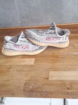 Yeezy Boost 350 Zebra sneakers - Snygga Yeezy Boost 350 Zebra sneakers med svartvitt mönster och röd text på sidan. Skorna har en krämfärgad, räfflad sula och är tillverkade i ett stickat material som ger en skön och flexibel känsla. Perfekta för dig som gillar streetwear och vill sticka ut.