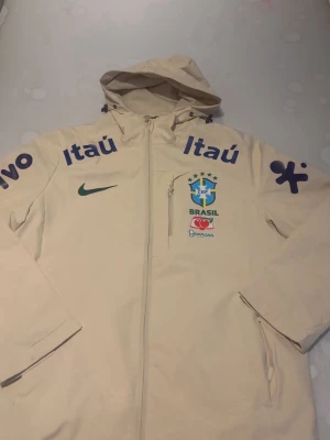 Beige Brasil Nike vindjacka med huva - Snygg beige vindjacka med huva från Nike, prydd med Brasilien fotbollslandslagets logga och flera sponsorloggor som Itaú och Guaraná. Jackan har dragkedja framtill, bröstficka med dragkedja och tryck i blått och grönt. Perfekt för dig som gillar sportig stil.
