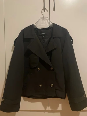 Svart dubbelknäppt kort rock - Storlek xl men sitter som en S! Snygg svart kort rock med dubbelknäppning och bred krage. Rocken har dekorativa knappar på ärmsluten och axelklaffar för en klassisk look. Passar perfekt till höst och vinter och ger en stilren vibe till din outfit.