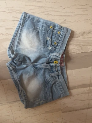 Ljusblå denimshorts med broderi - Säljer ett par ljusblå denimshorts med slitna detaljer och gula knappar. Shortsen har broderat mönster på ena bakfickan och en rosa etikett i midjan. Klassisk femficksmodell med bälteshällor och tvättad look.