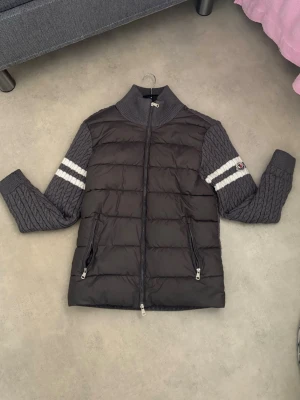 Grå dunväst från Moncler - Snygg grå dunväst från Moncler med stickade ärmar och två vita ränder på varje ärm. Västen har dragkedja framtill, två fickor med dragkedja och ribbad krage. Ärmarna är kabelstickade och har Moncler-logga på vänster sida.Priset kan diskuteras 🍾