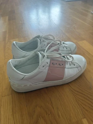 Vita sneakers med rosa detalj - Snygga vita sneakers med bred rosa rem över sidan och coola nitar runt hälen. Skorna har vita snören och en chunky sula. Tillverkade i slätt läderliknande material och har en modern, stilren look som passar till det mesta.