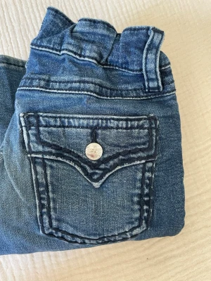 Blå bootcut jeans med fickdetalj - Säljer ett par blå bootcut jeans med snygga slitningar och en unik ficka bak med knappdetalj. Jeansen har klassisk femficksmodell och markerade sömmar. Perfekta för dig som gillar en avslappnad men trendig look. Litet hål mellan bakfickorna men går nog lätt att laga.
