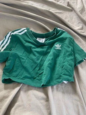 Grön croppad t-shirt från Adidas - Snygg grön croppad t-shirt från Adidas med klassiska vita ränder på ärmarna och Adidas-logga på bröstet. T-shirten har korta ärmar och rund halsringning. Perfekt för en sportig och avslappnad stil.