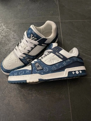 Louis Vuitton blåvita sneakers - Snygga Louis Vuitton sneakers i blått och vitt med klassiskt monogrammönster. Skorna har vita snören, detaljer i mocka och läder samt en robust sula. Perfekt för dig som vill sticka ut med exklusiv streetwear och premiumkänsla.