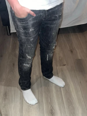 Svarta slitna jeans med hål - Säljer ett par svarta jeans med slitningar och hål på benen samt vita färgstänk för en riktigt edgy look. Jeansen har smal passform och klassisk femficksdesign. Perfekta för dig som gillar streetstyle och vill sticka ut lite extra. De är i ett väldigt bra skick. Hör av er vid funderingar.