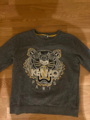 Grå sweatshirt med tiger från Kenzo - Snygg grå sweatshirt från Kenzo med det ikoniska tigerhuvudet broderat framtill i vitt, svart och gult. Rund halsringning och ribbade muddar. Perfekt för dig som vill ha en statementtröja med streetkänsla.