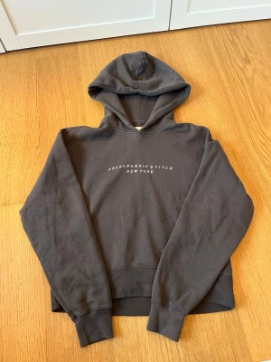 Mörkgrå hoodie Abercrombie & Fitch - Mysig mörkgrå hoodie från Abercrombie & Fitch med tryckt logga på bröstet. Tillverkad i mjukt material från Soft AF Collection och har en klassisk huva samt ribbade muddar vid ärmslut och nederkant. Perfekt för en avslappnad och stilren look.