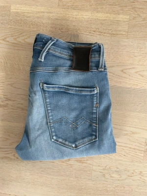 Replay anbass jeans - Säljer ett par ljusblå jeans i slim fit och klassisk femficksdesign. Jeansen har kontrastsömmar och en snygg detalj på bakfickorna. Materialet är denim med en lätt tvättad look och läderpatch i midjan. Finns en slitning och de har lagats i båda knäna vilket ger en ännu snyggare look.